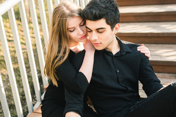 Beautiful young multiracial couple, student couple in love, sit wooden staircase in the city. Beautiful Turkish brunette guy hug a Caucasian girl in black clothing. Date young people in sunny weather