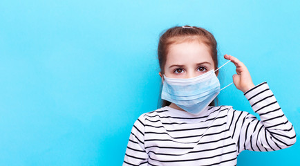 Child in a medical mask. COVID 19 world pandemic concept. Little girl in protective cloth disposable medical mask. Emotion baby photo. Copy space and blue background.