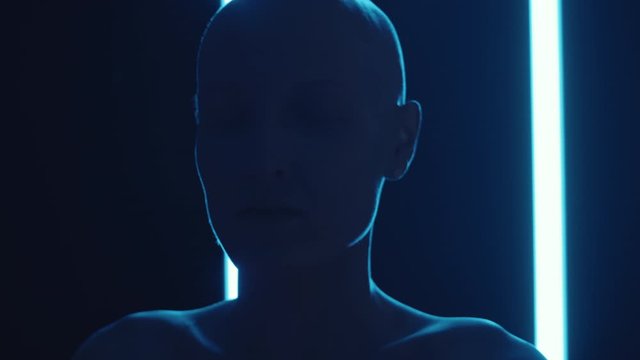 Handheld camera shot of young shirtless woman with bald head looking at camera while posing in dark studio with multicolored flickering neon light