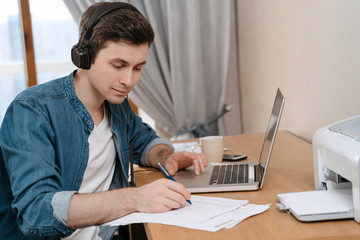 
Happy caucasian man focused on making notes while listening online webinar at home, wearing...