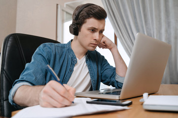 Serious young man in headphones sitting at desk in home office, working remotely, writting on papers, making notes, thinking on work task. Freelancer using laptop for work from home, preparing report