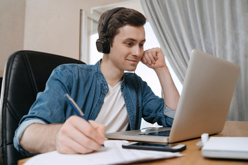 Happy guy student wearing headphones using laptop for distance education, enjoying conference video call, watching webinar, listening audio course, writing notes, studying with teacher, having lesson