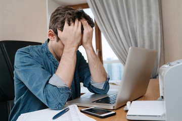 Tired stressed young man sitting in front of laptop, frustrated with problems or study failure, feel exhausted, having headache, upset with bad news, money problem, unsuccessful exam or test results
