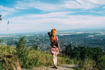 A young slender woman with red hair, dressed in a country-style dress, looks at the panorama of the city from the top of a mountain - 339362561