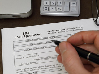 A Small Business Administration aka SBA loan application form, issued by the U.S.A. government, is shown up close, with a hand holding an ink pen about to fill out the paperwork.