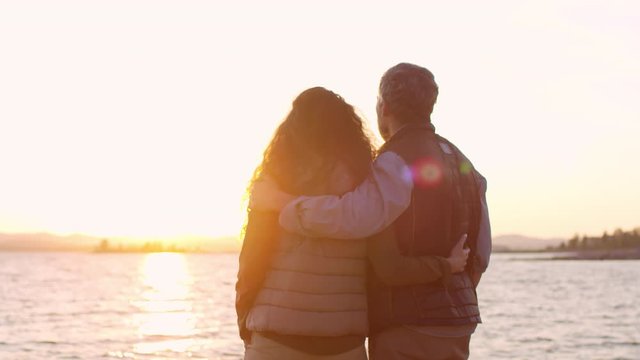 Rear view of romantic tourist couple embracing and speaking while standing on beach and looking at scenic sunset on the horizon over lake