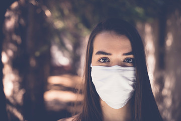 A young girl wearing a white mask for face protection against coronavirus in the woods. Looking into the camera. Blurry background. 