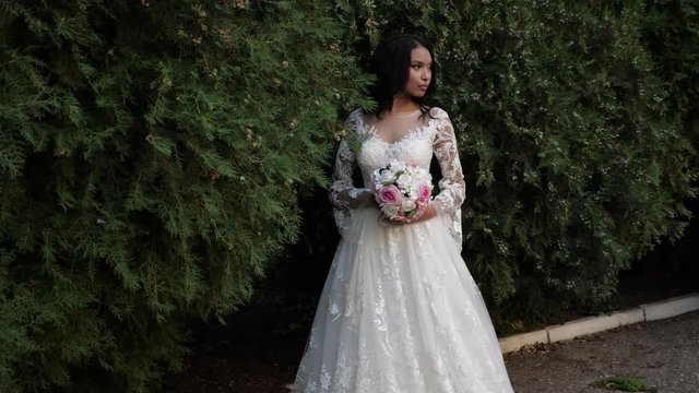 young bi-racial long haired woman in stylish wedding dress holds rose bouquet near bushes in green park slow motion