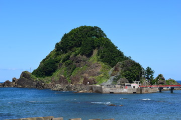 Obraz premium 白山島（東北の江ノ島）／ 山形県鶴岡市の由良海岸沖にある白山島は、「日本の渚百選」と、「快水浴場百選」に選ばれた由良海岸のシンボル的な島で、その景観から「東北の江ノ島」と呼ばれています。由良海岸から島までは赤い白山橋が架けられており、歩いて島に渡ることが出来ます。