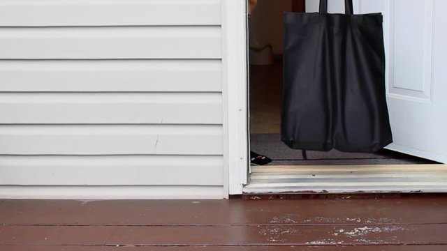 A Person Opening The Front Door Wearing Gloves, Picking Up A Groceries Bag From A Home Entrance