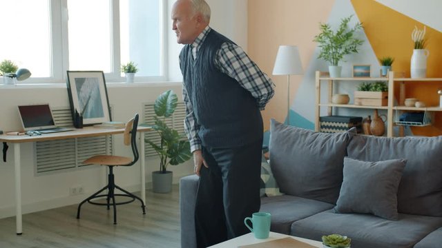 Slow motion of senior man standing up from sofa feeling backache touching back at home alone. Health problems, old people and retirement concept.