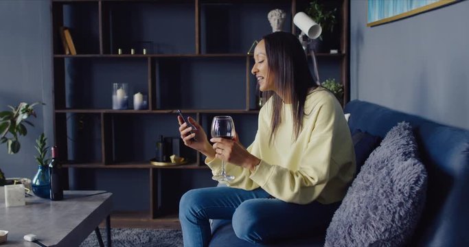 Attractive Hispanic female having a glass of wine while chatting with a friend or partner online in video chat, using a her phone at home