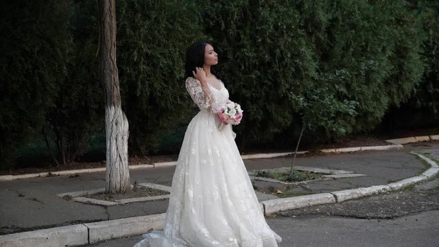 beautiful girl in long lacy wedding dress poses holding flower bouquet on city road by green park slow motion