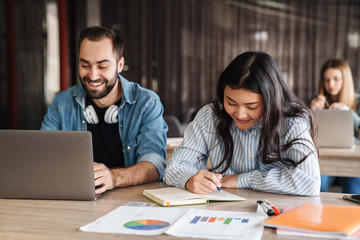 Photo of joyful students using laptop and writing in exercise book