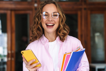 Photo of pleased woman using smartphone and holding exercise books