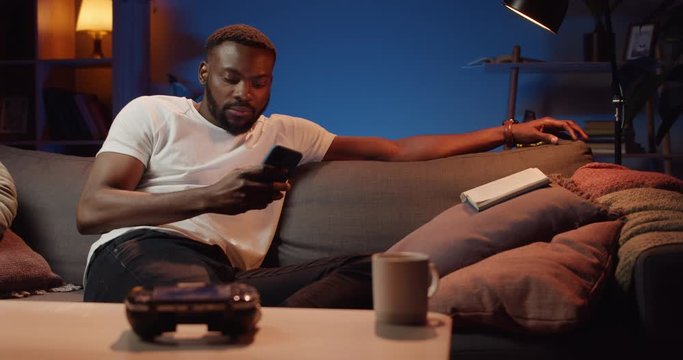 Front view of thoughtful young man using his smartphone while sitting on sofa. Bearded african guy scrolling phone screen while spending free time at home. Concept of leisure and tech