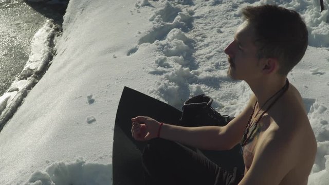 Man sitting on black yoga mat in snow with no shirt breathing heavily to prepare mind and body for frigid plunge into frozen mountain lake. Sun on shirtless guy hyperventilating before extreme sport.