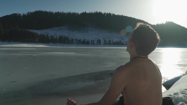 Young man with no shirt sitting beside frozen mountain lake in winter with snow on landscape and bright sunlight. Man hyperventilates to prepare his mind, lungs and body to go swimming in icy pond.