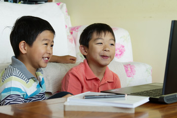 Two boys are using computer together for homework and education at home in the sitting room next to the sofa with some notebooks and a pencil next to them.Kids studying/E-learning from home concept.