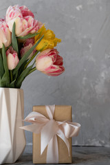 Colorful tulips in a white vase on the table on a light gray background, box with a gift. Kraft paper and bow. Mothers Day