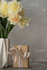Beautiful bouquet of yellow daffodils in a white vase on a gray table on a neutral background. Box with a gift on the table.