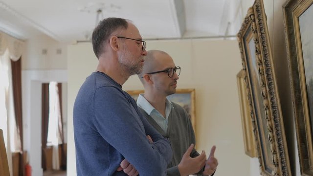 Two men in an art gallery look at a painting and talk about art