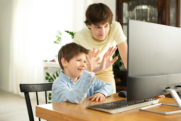 Smiling boy and teenager chatting online and waving at the computer screen. Quarantine and self-isolation due to coronavirus.