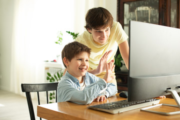 Smiling boy and teenager chatting online and waving at the computer screen. Quarantine and self-isolation due to coronavirus.