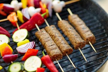 cooking, barbecue and food concept - close up of shish kebab meat and vegetables on bamboo skewers roasting to hot brazier grill outdoors