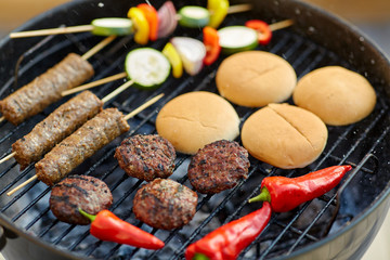 cooking, barbecue and food concept - close up of shish kebab meat, vegetables on bamboo skewers, burger buns and cutlets roasting on brazier grill