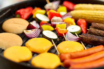 cooking, barbecue and food concept - close up of shish kebab meat, vegetables on bamboo skewers, burger buns with corn and sausages roasting on brazier grill outdoors