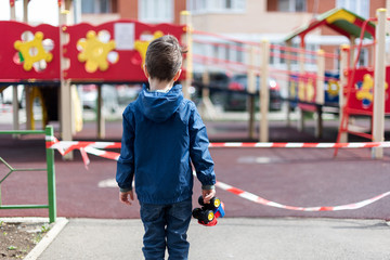 A child stands over a playground fenced with red ribbon, holds a toy car in his hand. Self isolation. It is forbidden to walk in the playgrounds