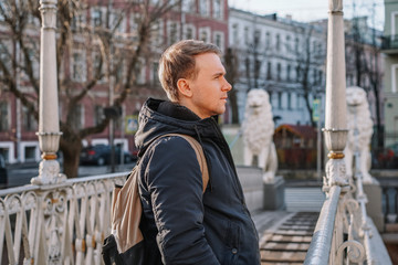 Fototapeta na wymiar A young blond man in a jacket and with a backpack stands on the bridge with statues of white lions in the middle of the canals of St. Petersburg, Attractions, morning spring