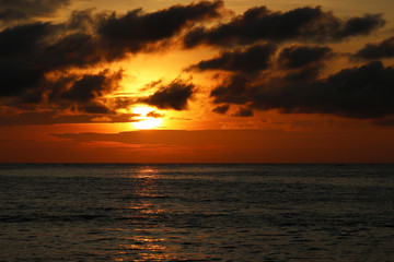 Fototapeta premium Sunset over the sea, orange sun is reflected in dramatic waves. Colorful sky with dark clouds, romantic background for travel and vacations