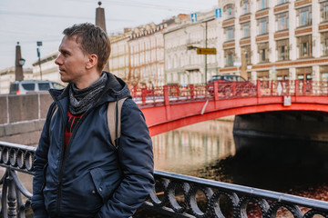 A blond man in a jacket and a sweater in the middle of the street against the background of the Sights of St. Petersburg, a beautiful historical building near the red bridge near the river channel, mo