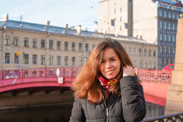 Fototapeta premium A girl with long hair in the middle of the street against the background of the Sights of St. Petersburg, a beautiful historical building near the red bridge near the river channel, morning spring