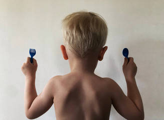 The boy stands with his back to the wall with a fork and spoon.