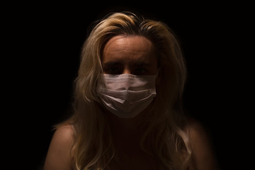 Blonde woman with white medical mask, sitting alone in the dark, looking sad and depressed