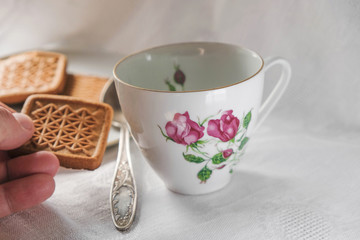 Vintage porcelain cup with spoon on a white background in Ukraine. Old woman's fingers are holding cookies in her hand. Morning light.