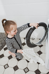 Cute girl in striped clothes puts things into the washing machine. Cleanliness and Household Concept