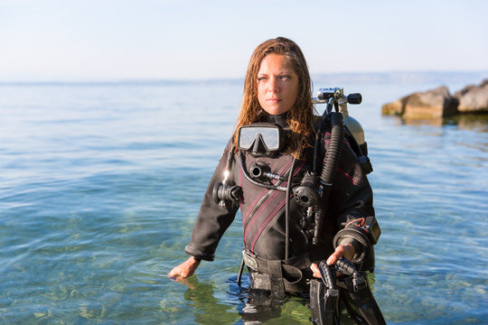 Female Scuba Diving Instructor Standing In Water Wearing A Dry Suit, A Twin Tank And Holding Fins