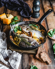 baked fish in cast iron skillet with mushrooms, herbs, cheese and spices on brown wooden background, nutritious dinner