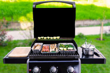 An opened gas grill with vegetables, meat, and sausages in aluminum barbeque trays or dip pans. The background with green grass, bushes, a sidewalk are blurred. copy space on the black grill cover. - obrazy, fototapety, plakaty