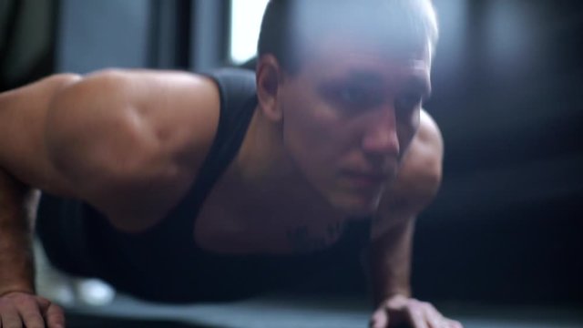 Close-up portrait of face of young man with muscular body doing push-ups exercise on the floor during sport workout training in modern dark gym. Concept of healthy lifestyle. Shooting in slow motion.