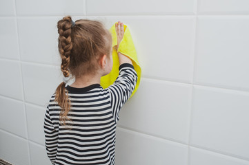 Cute girl in striped clothes washes a yellow rag on the wall. The child cleans at home. Household...