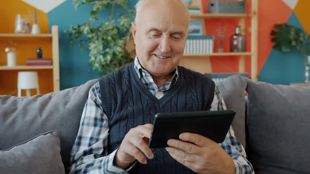 Cheerful senior man is swiping tablet screen enjoying online content sitting on couch at home. Modern technology, communication and old people concept.