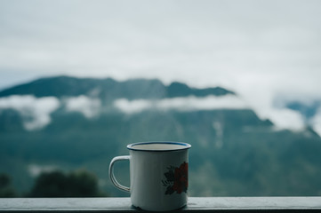 Close up view to traveler metal mug with hot tea on the balcony of hut in the mountains. Steam evaporating. Travel concept. Beverage during journey or hiking.