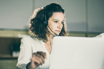 Focused mature call center operator looking at laptop. Serious curly woman working in office. Call...