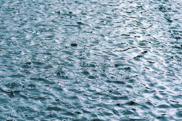 rain drops on the surface of water in a puddle with graduated shade of black shadow and reflection of blue sky
