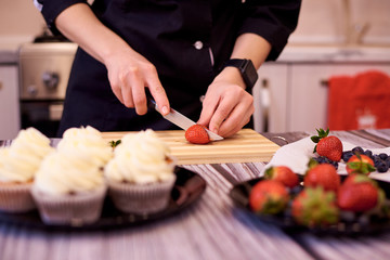 Obraz na płótnie Canvas Female hands with a knife cut strawberries on a cutting board on the kitchen table. Cooking cupcakes with strawberries and blueberries.Selective focus.